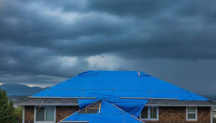 Blue emergency tarp secured over a damaged roof under a dark storm sky